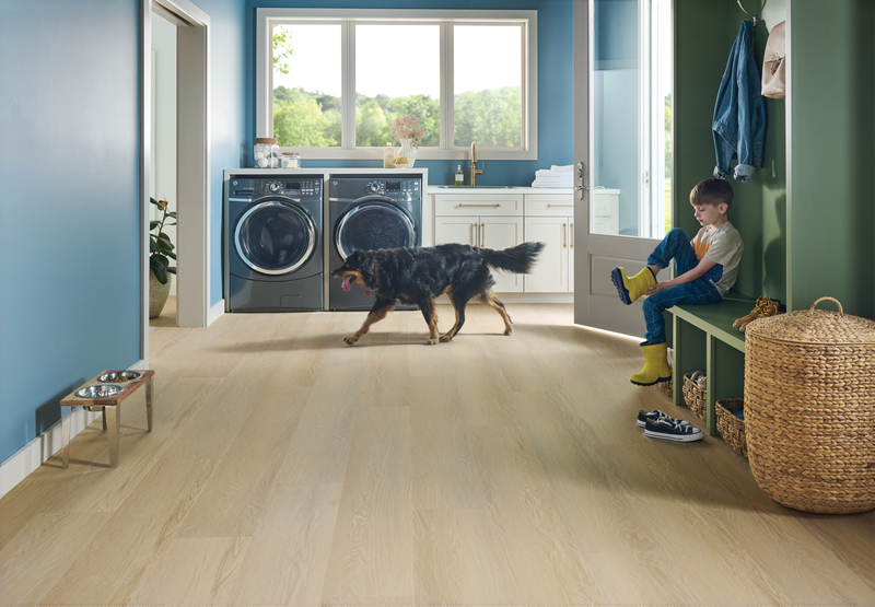 Mudroom area with a dog walking past a washer/dryer and child putting on boots to show the versatility of WPC flooring